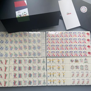 Puede incluir: Un juego de Mahjong con fichas blancas que presentan gr&aacute;ficos coloridos, incluyendo balones de f&uacute;tbol, banderas e ilustraciones deportivas. El conjunto incluye una caja de almacenamiento negra, una tarjeta blanca y un sobre negro con un logo.