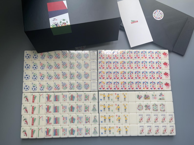 Puede incluir: Un juego de Mahjong con fichas blancas que presentan gr&aacute;ficos coloridos, incluyendo balones de f&uacute;tbol, banderas e ilustraciones deportivas. El conjunto incluye una caja de almacenamiento negra, una tarjeta blanca y un sobre negro con un logo.
