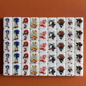 Puede incluir: Un juego de 48 domin&oacute;s blancos con im&aacute;genes de personajes de Sonic the Hedgehog. Los personajes incluyen Sonic, Tails, Knuckles, Amy, Shadow y Eggman. Los domin&oacute;s est&aacute;n dispuestos en una cuadr&iacute;cula.