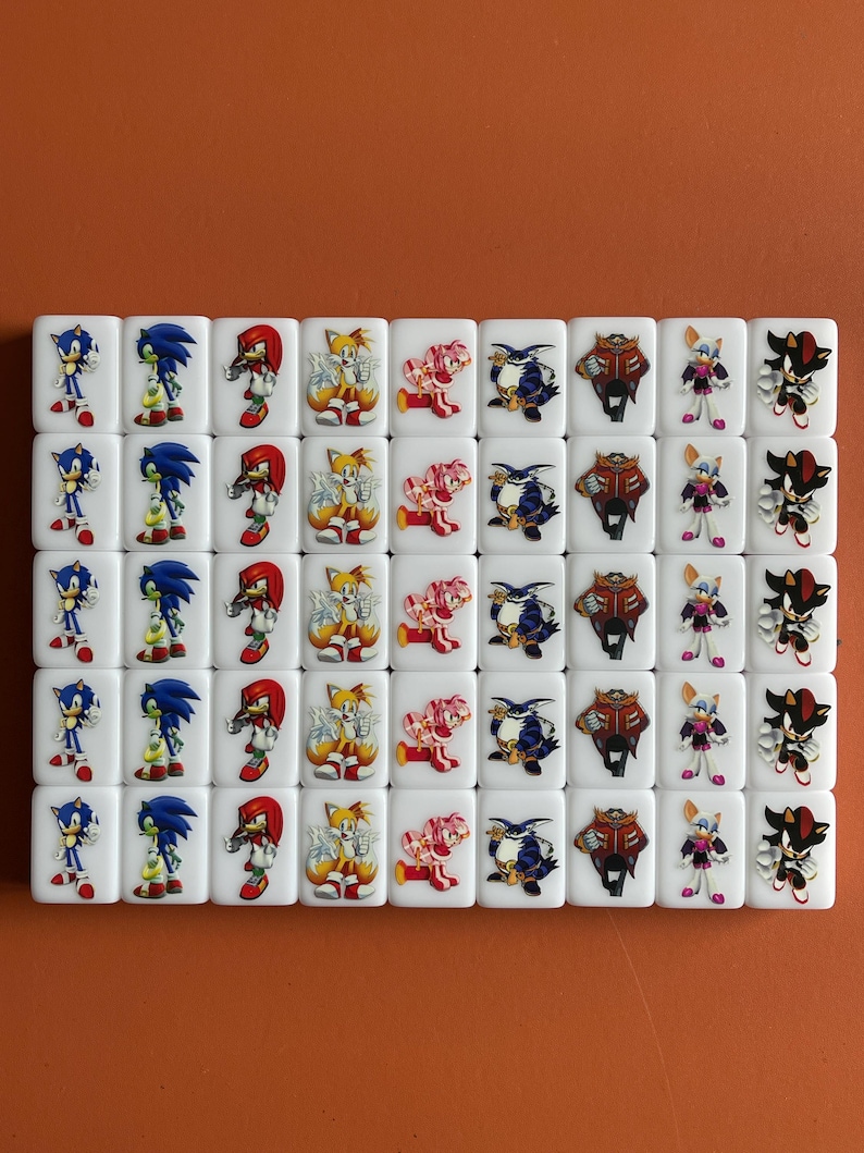 Puede incluir: Un juego de 48 domin&oacute;s blancos con im&aacute;genes de personajes de Sonic the Hedgehog. Los personajes incluyen Sonic, Tails, Knuckles, Amy, Shadow y Eggman. Los domin&oacute;s est&aacute;n dispuestos en una cuadr&iacute;cula.