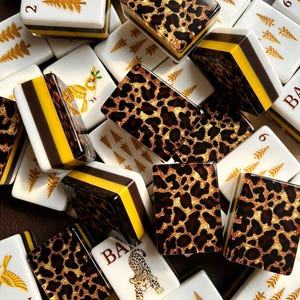 May include: A collection of rectangular mahjong tiles featuring a mix of white tiles with gold tree and bird illustrations, and tiles with a brown, black, and gold leopard print pattern. Some tiles display the text BAM and numerical markings.