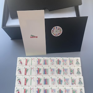 Puede incluir: Un juego de fichas de mahjong blancas dispuestas en cuatro filas, con ilustraciones coloridas de p&aacute;jaros, banderas y motivos deportivos numerados del uno al nueve. Incluye una caja negra, una tarjeta blanca y un sobre negro con pegatina.