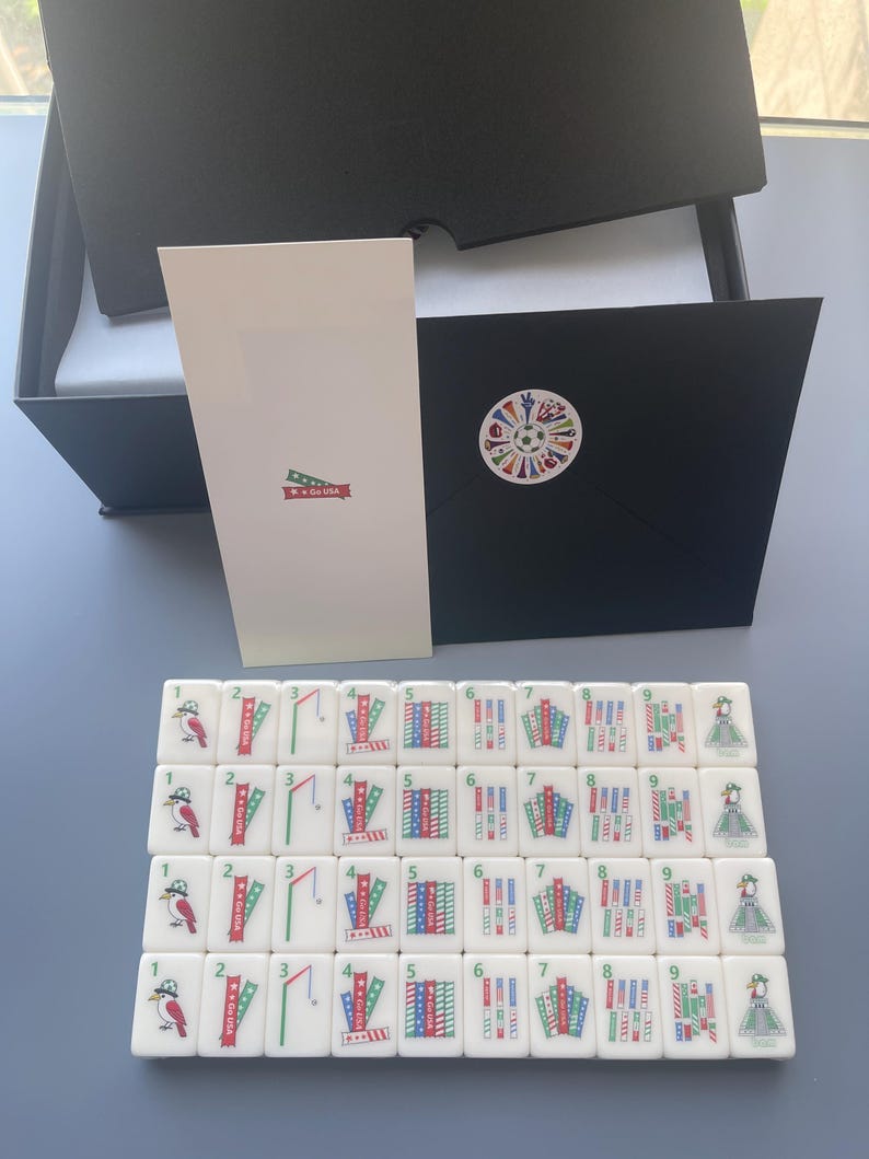 Puede incluir: Un juego de fichas de mahjong blancas dispuestas en cuatro filas, con ilustraciones coloridas de p&aacute;jaros, banderas y motivos deportivos numerados del uno al nueve. Incluye una caja negra, una tarjeta blanca y un sobre negro con pegatina.