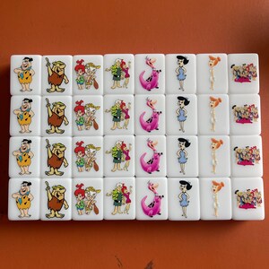 May include: A set of white tiles featuring cartoon characters from The Flintstones. Each tile displays a different character or group of characters in vibrant colors. The tiles are arranged in rows and columns.