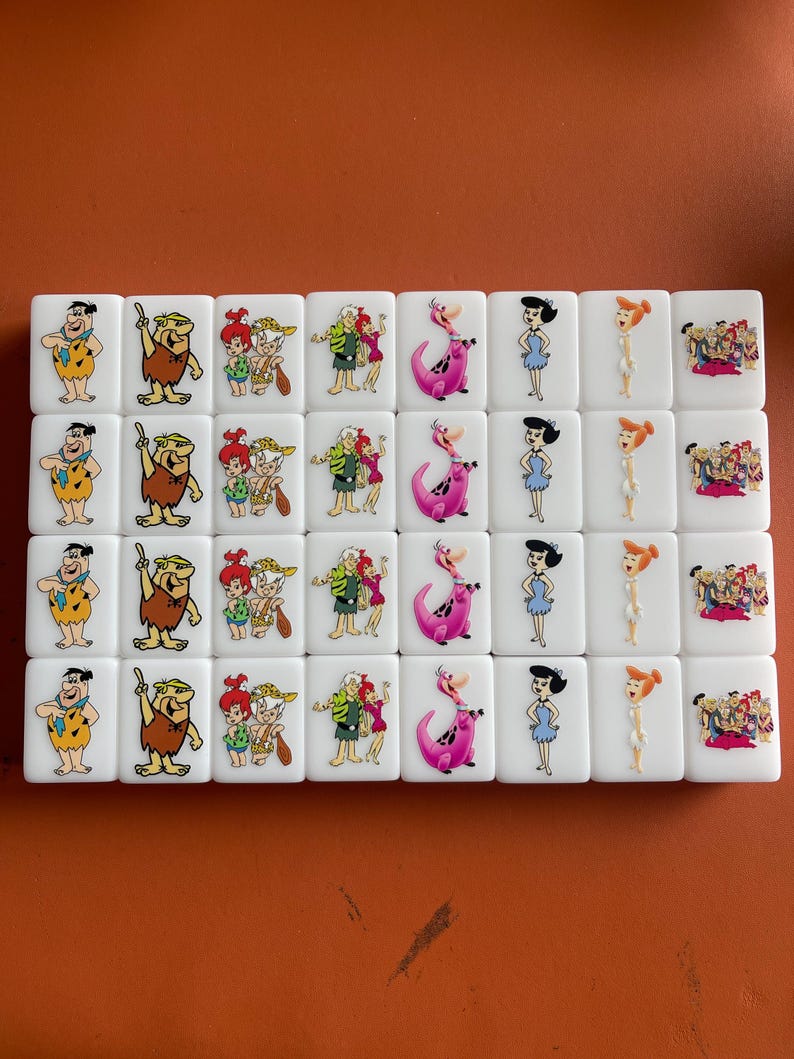 May include: A set of white tiles featuring cartoon characters from The Flintstones. Each tile displays a different character or group of characters in vibrant colors. The tiles are arranged in rows and columns.