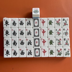 May include: A set of white Mahjong tiles with black and red symbols and characters. The tiles include directional markers, card symbols, and flower tiles. A single Joker tile is at the top. The tiles are arranged on an orange surface.
