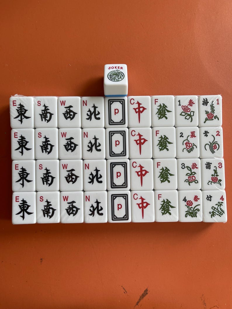 May include: A set of white Mahjong tiles with black and red symbols and characters. The tiles include directional markers, card symbols, and flower tiles. A single Joker tile is at the top. The tiles are arranged on an orange surface.