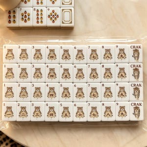 May include: A set of white rectangular mahjong tiles arranged in rows, featuring illustrations of leopards holding circular tokens with Chinese numerals one through nine and the word CRAK. The tiles are encased in clear plastic packaging.