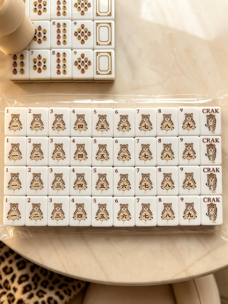 May include: A set of white rectangular mahjong tiles arranged in rows, featuring illustrations of leopards holding circular tokens with Chinese numerals one through nine and the word CRAK. The tiles are encased in clear plastic packaging.