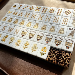 May include: A set of white Mahjong tiles arranged in a grid, featuring gold-colored illustrations of flowers, trees, numbers, and leopard faces. Several tiles are labeled with the word JOKER, CRAK, or BAM, and two tiles feature a brown leopard print pattern.