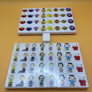 May include: Two sets of white tiles, likely for a game, are arranged on a yellow surface. The top set features Sesame Street characters, while the bottom set showcases Peanuts characters. A single white tile sits between the sets.