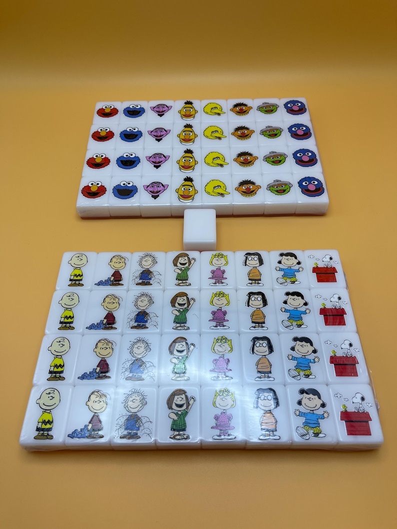 May include: Two sets of white tiles, likely for a game, are arranged on a yellow surface. The top set features Sesame Street characters, while the bottom set showcases Peanuts characters. A single white tile sits between the sets.