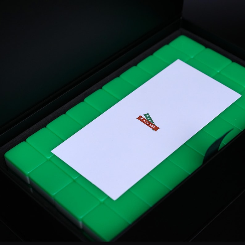 Puede incluir: Una caja de almacenamiento negra que contiene una cuadr&iacute;cula de azulejos rectangulares de color verde brillante. Una tarjeta rectangular blanca descansa sobre los azulejos, con un peque&ntilde;o gr&aacute;fico de dos cintas cruzadas en rojo y verde con el texto Go USA.
