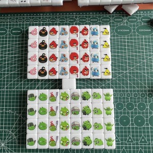 May include: A set of 36 white ceramic dominoes with Angry Birds characters printed on them. The dominoes are arranged in a square pattern on a green cutting mat. The dominoes are 3.8 cm x 3.8 cm.