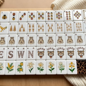 May include: A set of white mahjong tiles arranged in a grid on a wooden surface. The tiles feature various illustrations including ladybugs, pine trees, leopards, flowers, and the text E, S, W, N, CRAK, BAM, and JOKER. Two tiles have a leopard print pattern.