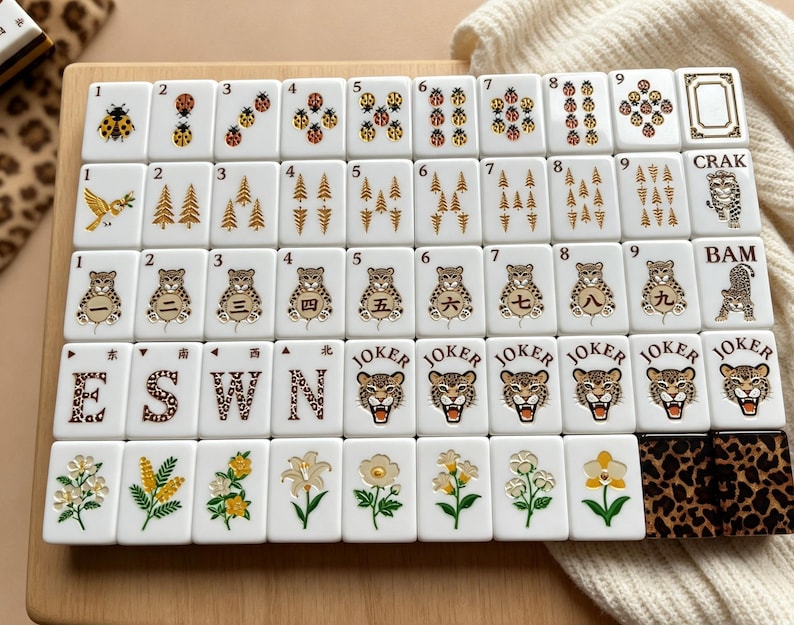 May include: A set of white mahjong tiles arranged in a grid on a wooden surface. The tiles feature various illustrations including ladybugs, pine trees, leopards, flowers, and the text E, S, W, N, CRAK, BAM, and JOKER. Two tiles have a leopard print pattern.