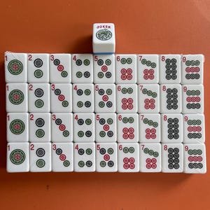 May include: A set of white Mahjong tiles with red, green, and black markings, arranged on an orange surface. The tiles are numbered 1 through 9, with various suit symbols. A single tile marked "JOKER" is also present.