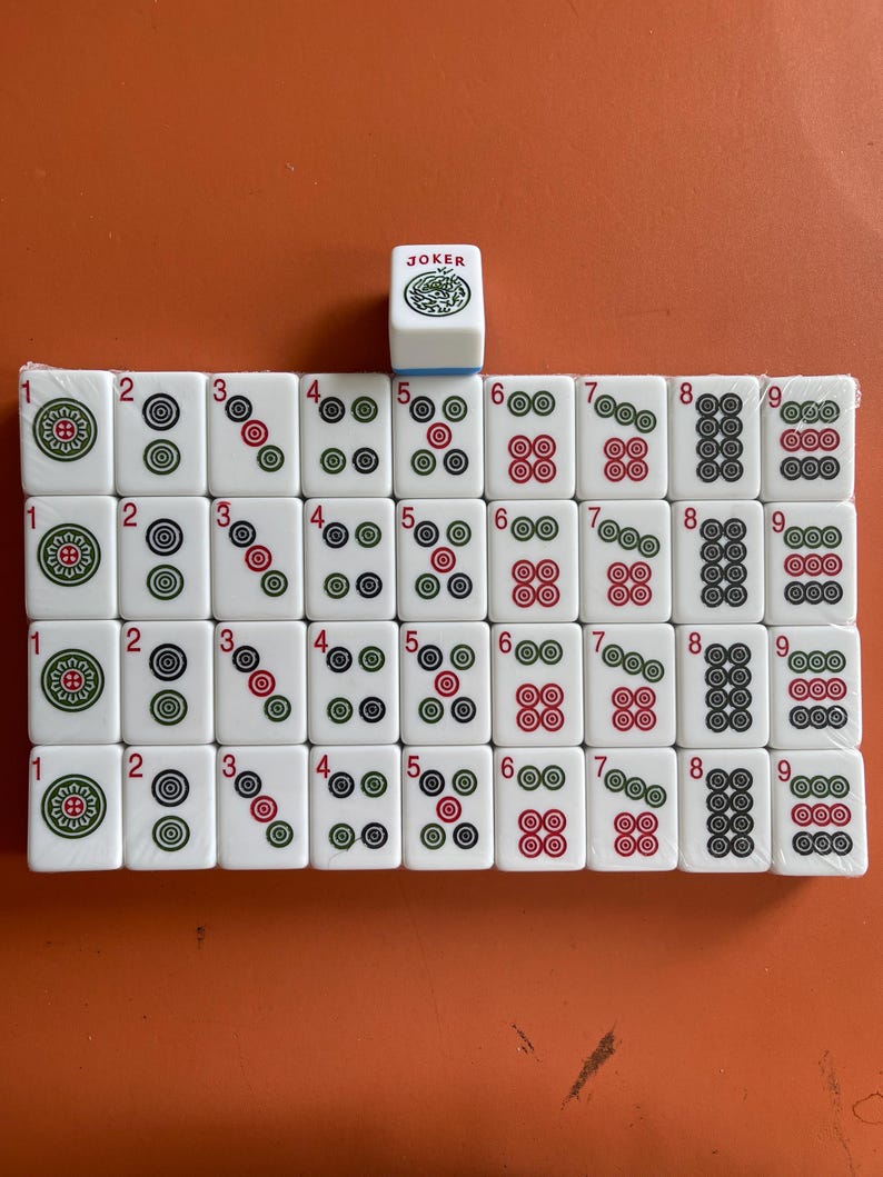 May include: A set of white Mahjong tiles with red, green, and black markings, arranged on an orange surface. The tiles are numbered 1 through 9, with various suit symbols. A single tile marked "JOKER" is also present.