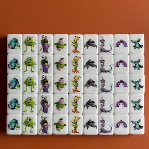 Puede incluir: Un conjunto de 36 dados blancos con diferentes personajes de dibujos animados de las pel&iacute;culas de Disney Pixar. Los personajes incluyen Mike Wazowski, Sulley, Boo, Randall y la puerta de Monstruos S.A. Los dados est&aacute;n dispuestos en seis filas de seis dados cada una.