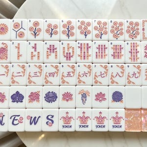 Puede incluir: Un juego de fichas de Mahjong blancas con diseños grabados en rosa, morado y naranja. Las fichas muestran motivos florales, tallos de bambú, números chinos, la palabra NEWS, iconos de Joker y dos fichas con un acabado iridiscente brillante.