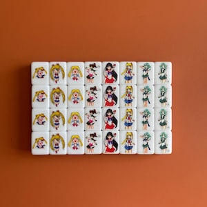 May include: A set of 24 white square tiles with images of anime characters. The characters are dressed in sailor outfits and have different colored hair and eyes. The tiles are arranged in a rectangular grid.
