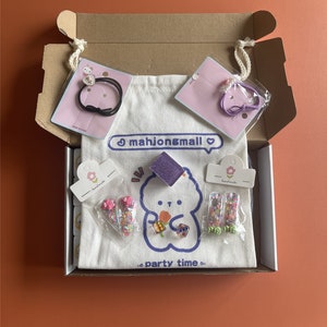 May include: A cardboard box filled with various accessories, including hair ties, a tote bag, and decorative items. The tote bag features a cartoon bear design and the text "mahjong&mall" and "party time". The accessories are packaged in clear plastic.