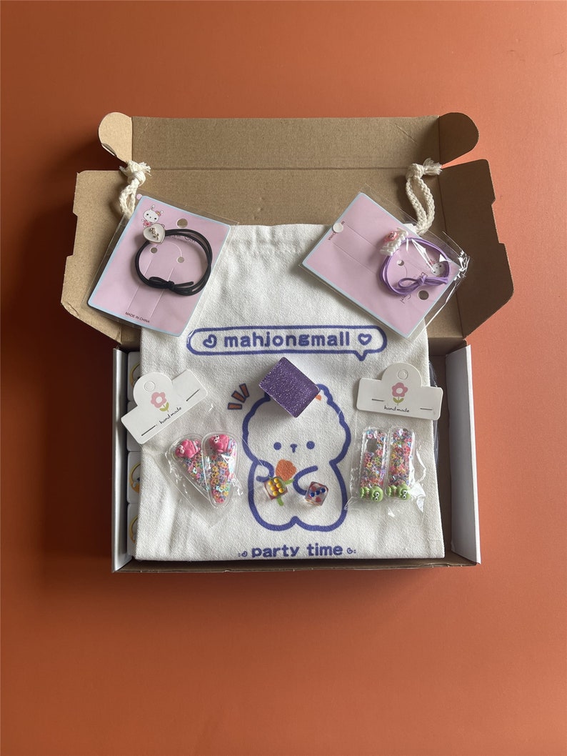 May include: A cardboard box filled with various accessories, including hair ties, a tote bag, and decorative items. The tote bag features a cartoon bear design and the text "mahjong&mall" and "party time". The accessories are packaged in clear plastic.