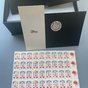 Puede incluir: Un conjunto de fichas de Mahjong blancas dispuestas en cuatro filas, cada una con caracteres chinos rojos y un peque&ntilde;o gr&aacute;fico de un bal&oacute;n de f&uacute;tbol con una hoja de arce roja. Incluye una tarjeta blanca alta y un sobre negro con una pegatina.