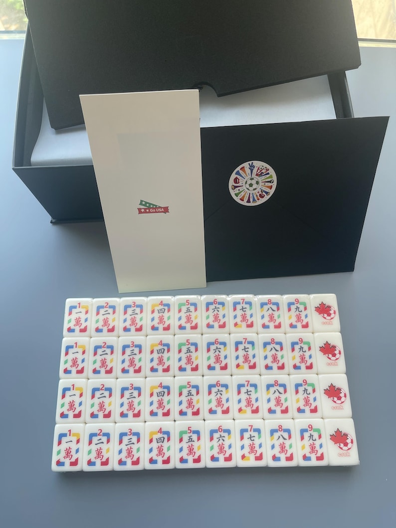Puede incluir: Un conjunto de fichas de Mahjong blancas dispuestas en cuatro filas, cada una con caracteres chinos rojos y un peque&ntilde;o gr&aacute;fico de un bal&oacute;n de f&uacute;tbol con una hoja de arce roja. Incluye una tarjeta blanca alta y un sobre negro con una pegatina.