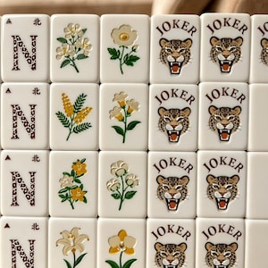 May include: A set of rectangular white mahjong tiles arranged in a grid. The tiles feature various designs including the letter N with a leopard print pattern, diverse floral illustrations in green, yellow, and white, and a leopard face with the text JOKER.