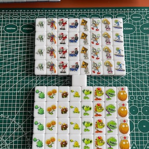 May include: A set of 38 white dominoes featuring colourful images of plants and zombies from the popular video game Plants vs. Zombies. The dominoes are arranged in a grid pattern on a green grid surface. The dominoes are approximately 3.8 cm wide and 2.8 cm tall.