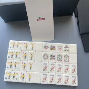 Puede incluir: Un conjunto de fichas de mahjong rectangulares blancas con gr&aacute;ficos coloridos de jugadores de f&uacute;tbol, se&ntilde;ales de &aacute;rbitro y el texto "We Are 26". Una tarjeta blanca alta con un peque&ntilde;o gr&aacute;fico de "Go USA" se encuentra detr&aacute;s de las fichas.