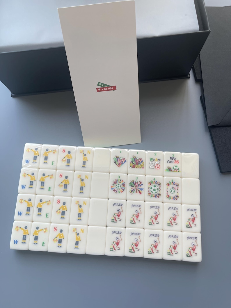 Puede incluir: Un conjunto de fichas de mahjong rectangulares blancas con gr&aacute;ficos coloridos de jugadores de f&uacute;tbol, se&ntilde;ales de &aacute;rbitro y el texto "We Are 26". Una tarjeta blanca alta con un peque&ntilde;o gr&aacute;fico de "Go USA" se encuentra detr&aacute;s de las fichas.