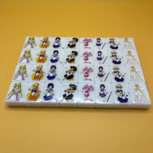 May include: A set of white mahjong tiles featuring colorful anime character illustrations. Each tile displays a different character in vibrant colors, set against a yellow background. The tiles are arranged in neat rows, showcasing the detailed artwork.
