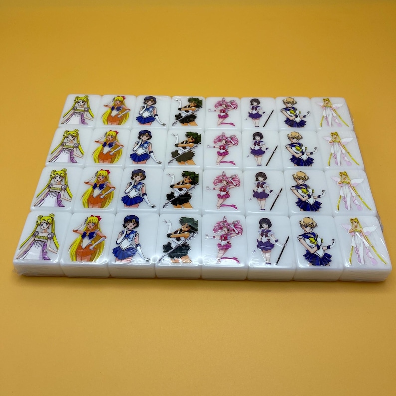 May include: A set of white mahjong tiles featuring colorful anime character illustrations. Each tile displays a different character in vibrant colors, set against a yellow background. The tiles are arranged in neat rows, showcasing the detailed artwork.