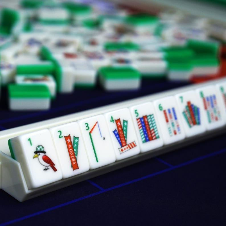 Puede incluir: Un conjunto de fichas de Mahjong blancas y verdes dispuestas en un soporte. La fila frontal muestra fichas numeradas del uno al ocho, con ilustraciones coloridas de un p&aacute;jaro, cintas con el texto Go USA, un bal&oacute;n de f&uacute;tbol y patrones de rayas.