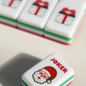 Op de afbeelding: Een rechthoekige witte spelsteen met een afbeelding van de Kerstman en de rode tekst JOKER, voorzien van groene en rode lagen aan de randen. Andere stenen op de achtergrond tonen afbeeldingen van cadeauverpakkingen in hetzelfde wit, groen en rood.