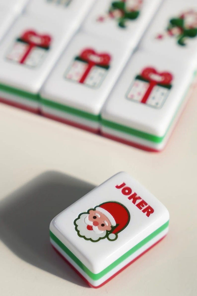 Op de afbeelding: Een rechthoekige witte spelsteen met een afbeelding van de Kerstman en de rode tekst JOKER, voorzien van groene en rode lagen aan de randen. Andere stenen op de achtergrond tonen afbeeldingen van cadeauverpakkingen in hetzelfde wit, groen en rood.