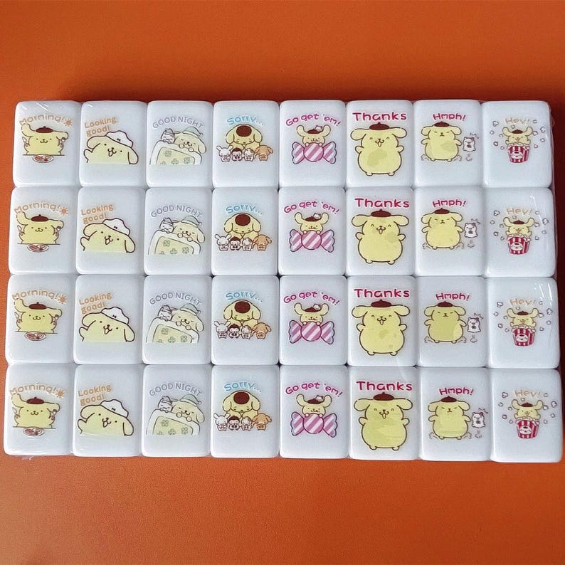 Op de afbeelding: Een set van 36 witte domino's met kleurrijke cartoonafbeeldingen van een hond met een hoed. De domino's bevatten zinnen zoals "Morning", "Looking Good", "Good Night", "Sorry", "Go get 'em", "Thanks", "Hmph!" en "He've".