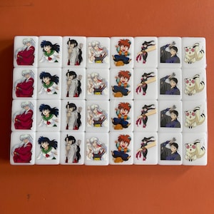 May include: A set of white Mahjong tiles featuring colorful anime character illustrations. Each tile displays a different character, with a variety of poses and outfits. The tiles are arranged in neat rows and columns, creating a visually appealing pattern.