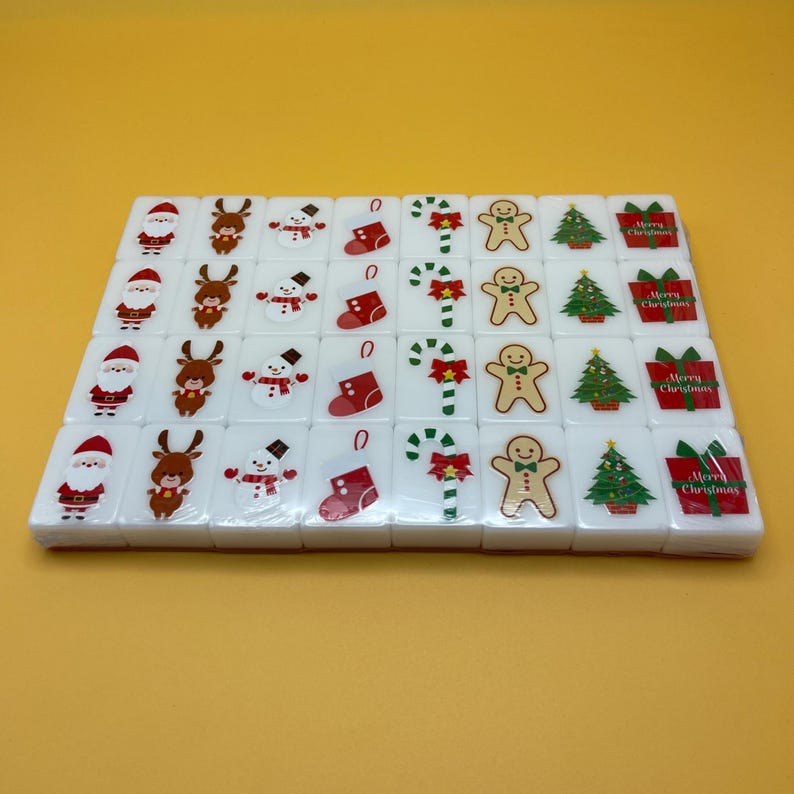 May include: A set of white Mahjong tiles featuring Christmas-themed illustrations. The tiles display images of Santa Claus, reindeer, snowmen, stockings, candy canes, gingerbread men, Christmas trees, and gift boxes. The tiles are arranged on a yellow surface.