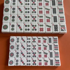 May include: Two sets of white Mahjong tiles with red, green, and black markings. The tiles are arranged in rows and columns, displaying numbers and symbols. The sets are on an orange surface.