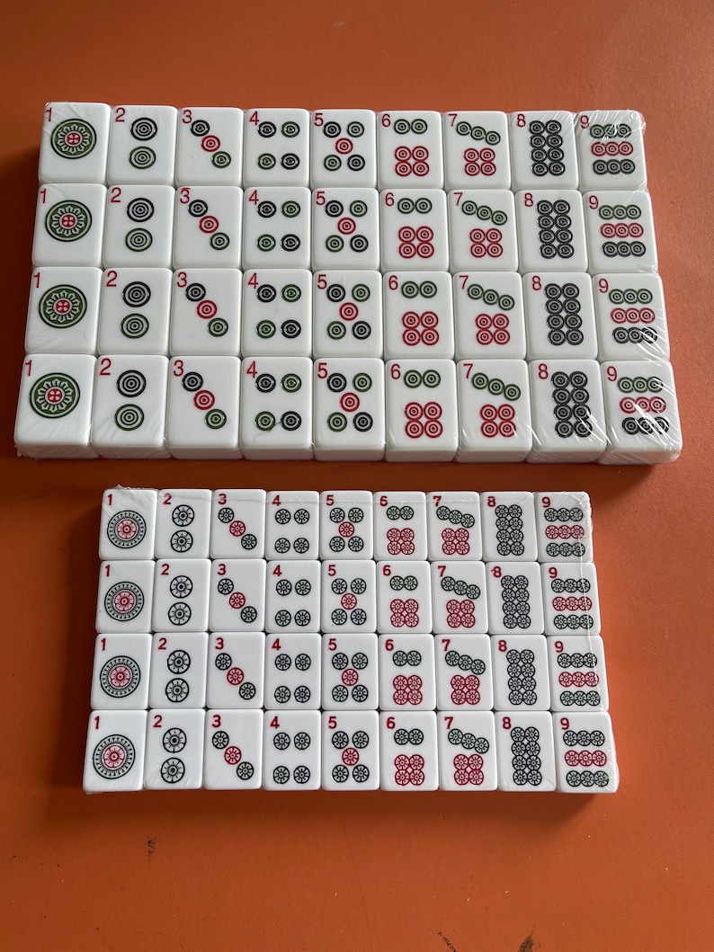May include: Two sets of white Mahjong tiles with red, green, and black markings. The tiles are arranged in rows and columns, displaying numbers and symbols. The sets are on an orange surface.