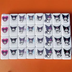 Puede incluir: Un conjunto de 36 fichas de cer&aacute;mica blanca con personajes de dibujos animados negros y morados. Los personajes tienen diferentes expresiones y est&aacute;n dispuestos en una cuadr&iacute;cula.