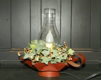 May include: A decorative electric candle lamp with a clear glass chimney. The lamp features a faux candle with a soft glow, surrounded by green ivy and berries, set in a rustic, dark red metal base. The lamp has a handle and a power cord.