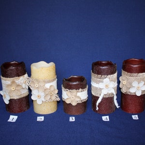 May include: Five decorative pillar candles with burlap and lace accents. The candles are various shades of brown and cream, adorned with white floral embellishments and wooden buttons. Each candle has a unique design.