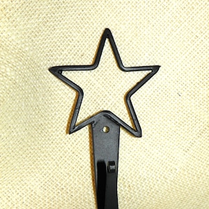 May include: Black metal star-shaped wall hook. The hook features a five-pointed star design at the top, with a curved hook extending downwards. The hook is mounted on a rectangular base with a small hole for hanging. The background is a textured, beige fabric.