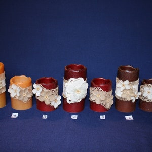 May include: A collection of seven pillar candles in varying heights and colors, including orange, red, and brown. Each candle is decorated with burlap, lace, and floral accents, creating a rustic and decorative aesthetic.