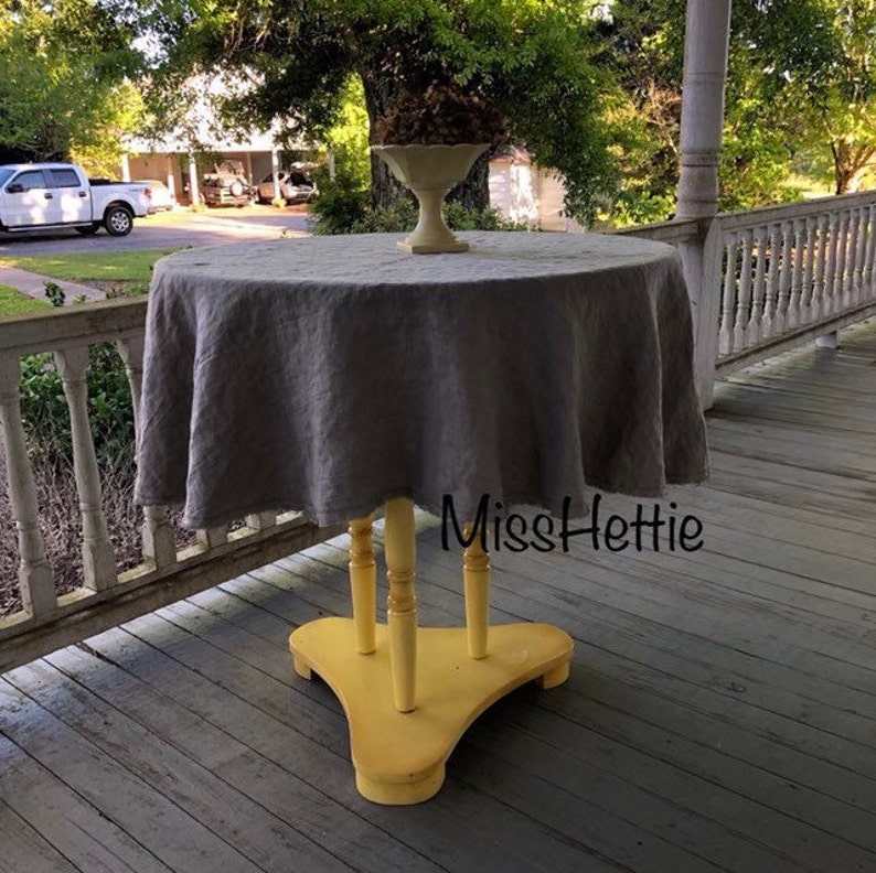 May include: A round gray linen tablecloth draped over a yellow painted table with three legs. The table is on a wooden porch.