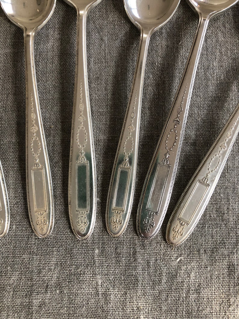 May include: Six silver spoons with ornate designs on the handles. The spoons have a rectangular space on the handle where a name or inscription could be engraved.